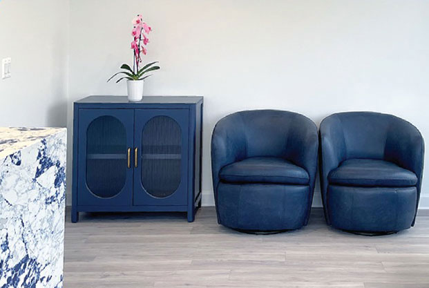 This is a photograph of an interior space featuring two blue armchairs with matching ottomans, a contemporary sideboard with a dark finish, a marble-topped console table, a modern blue cabinet, and a large abstract wall art. The room has a clean, minimalist aesthetic with white walls and a light wooden floor.