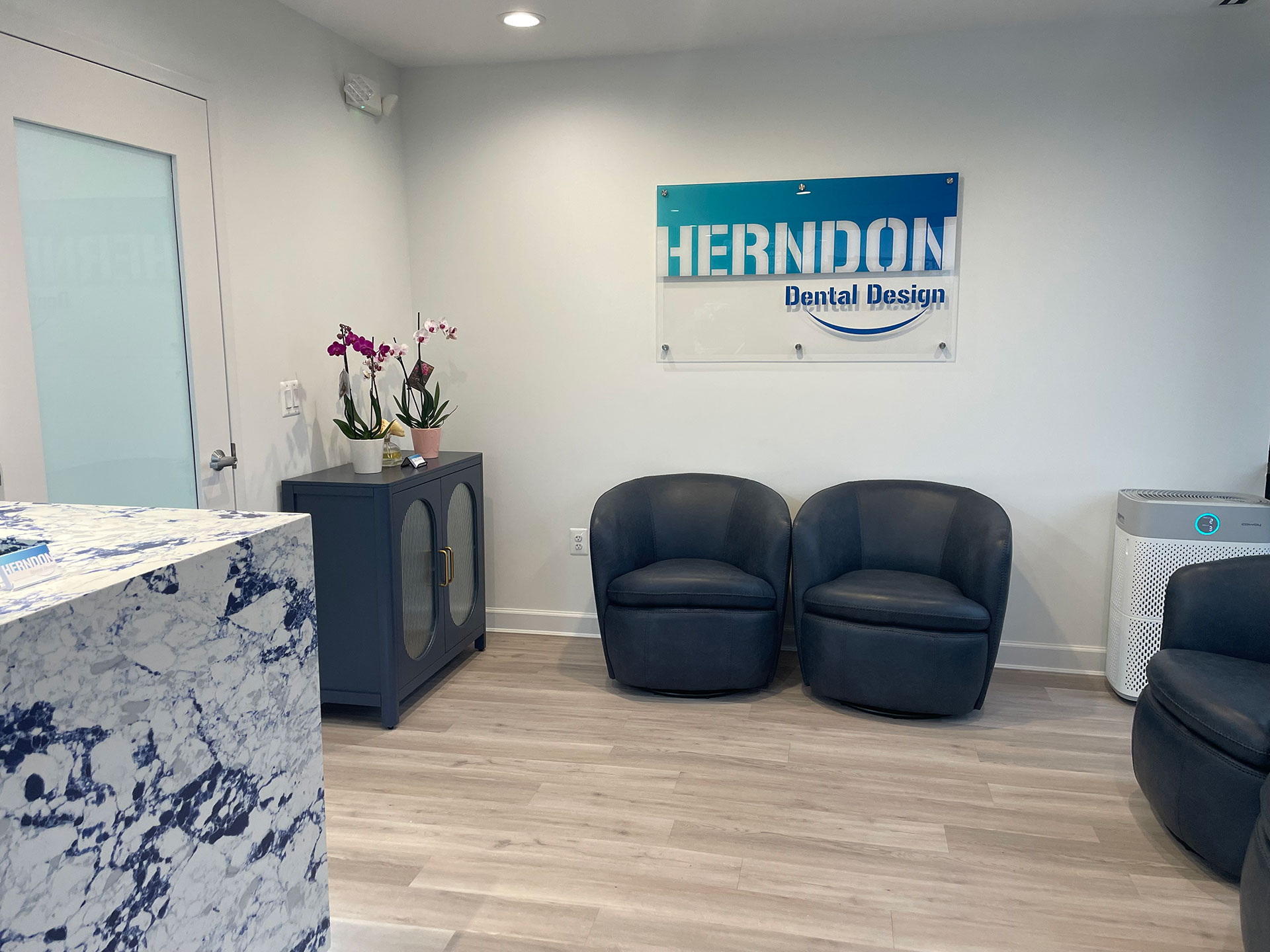 An interior office space with a modern reception area featuring a marble countertop, black leather chairs, a white sign with blue lettering that reads  Hermondon Dental Design,  and a large window letting in natural light.